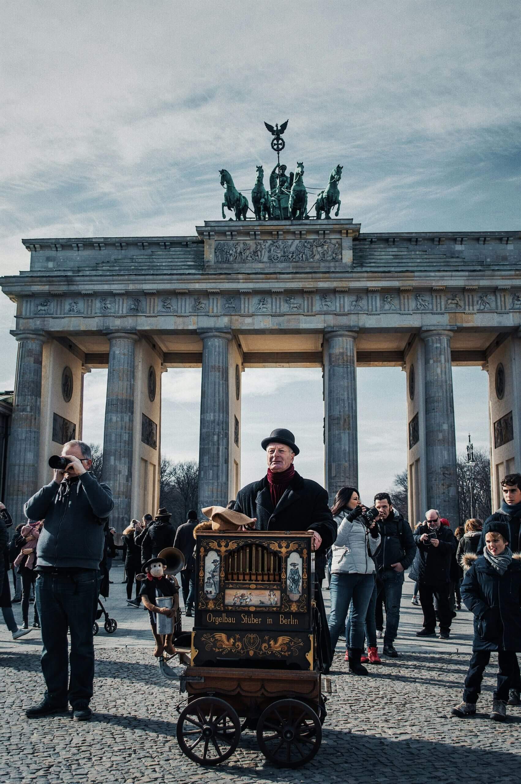 Man with bowler hat playing organ grinder in front of Berlin Brandenburg Gate with crowd around.
