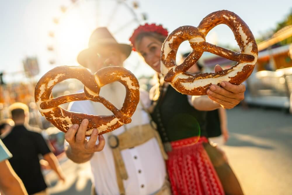 Man and woman in traditional Bavarian attire holding large pretzels at an outdoor fair.