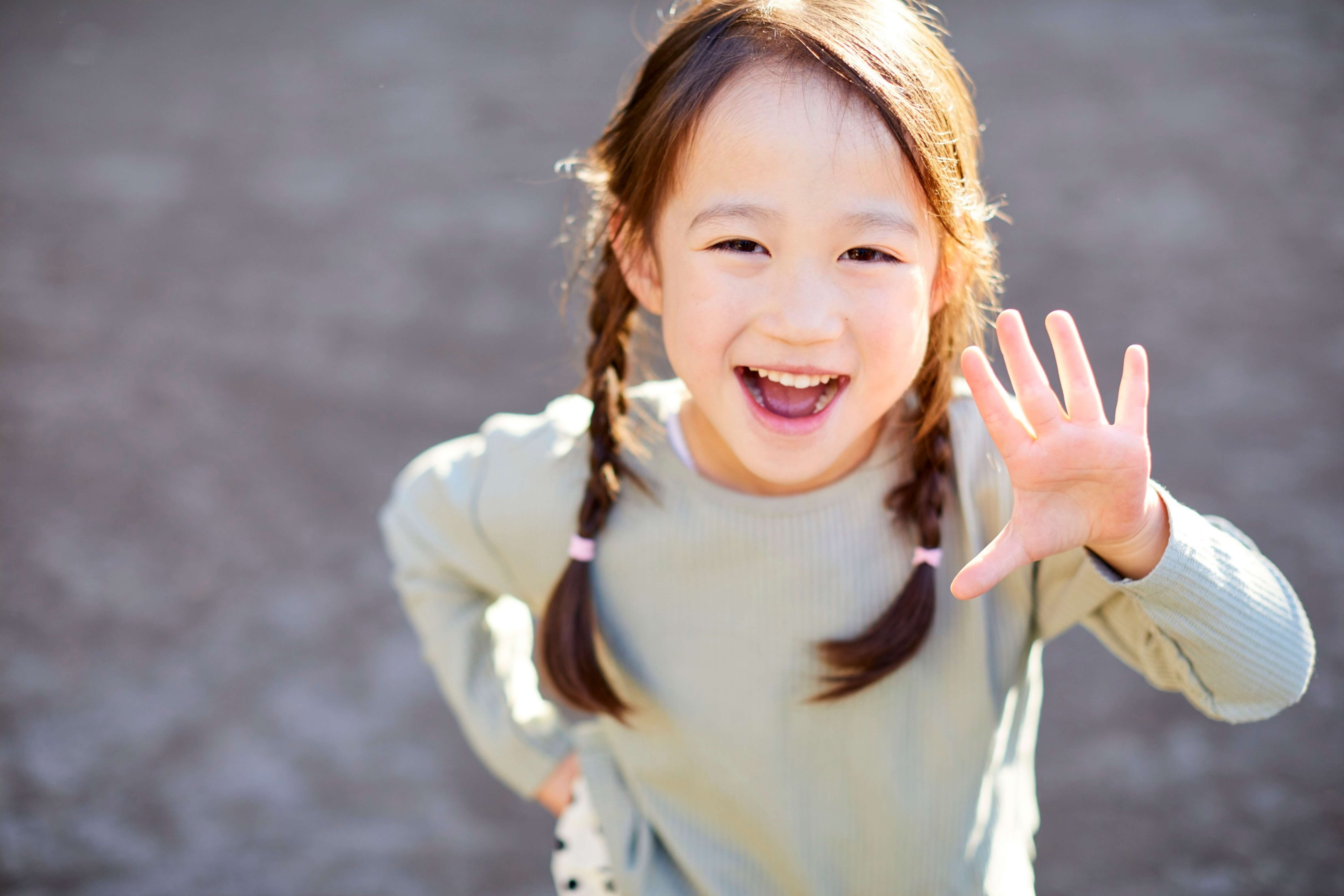 Smiling young girl with braided hair waving her hand outdoors with a blurred background.