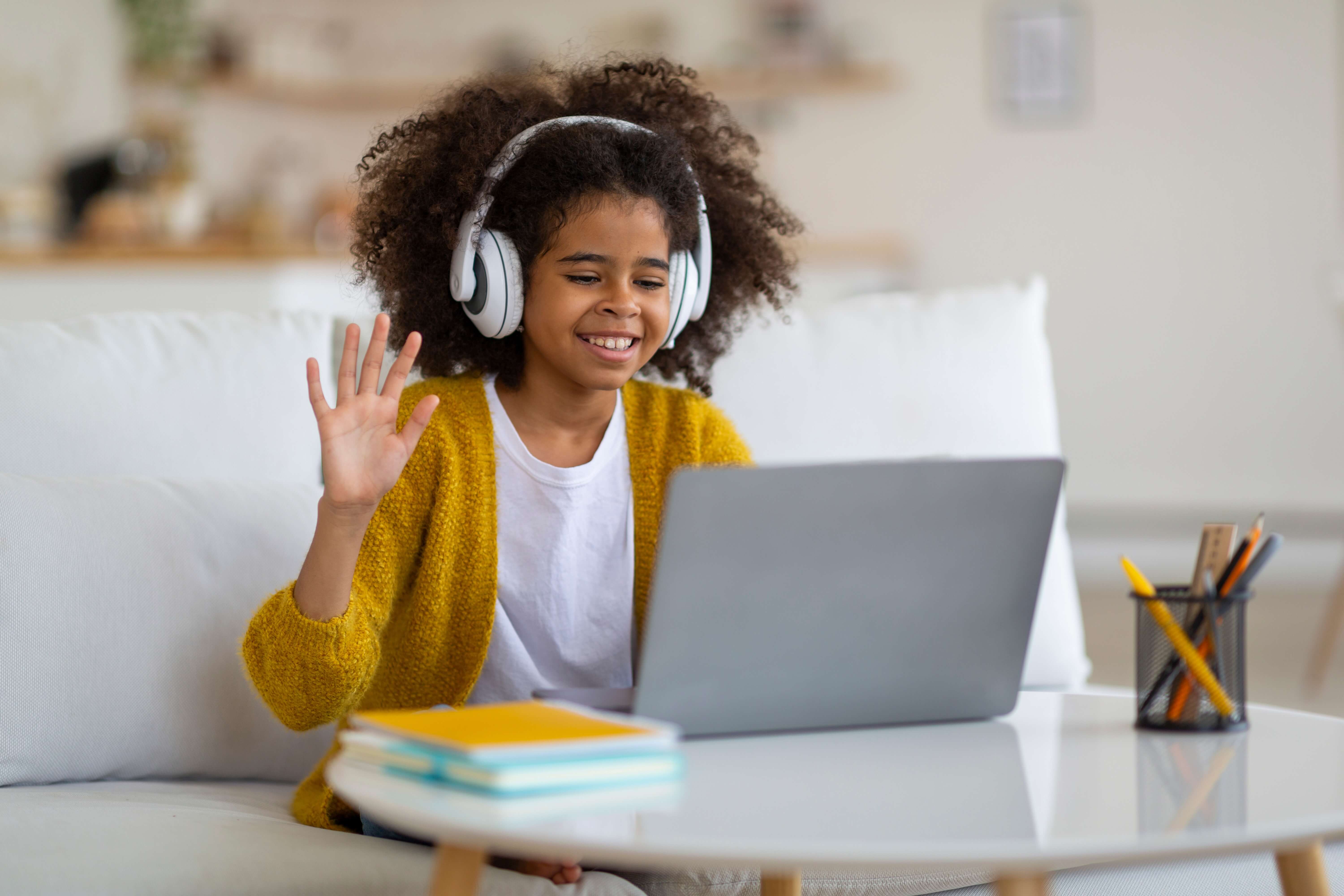 Smiling girl with headphones waving at a laptop during a video call at home.