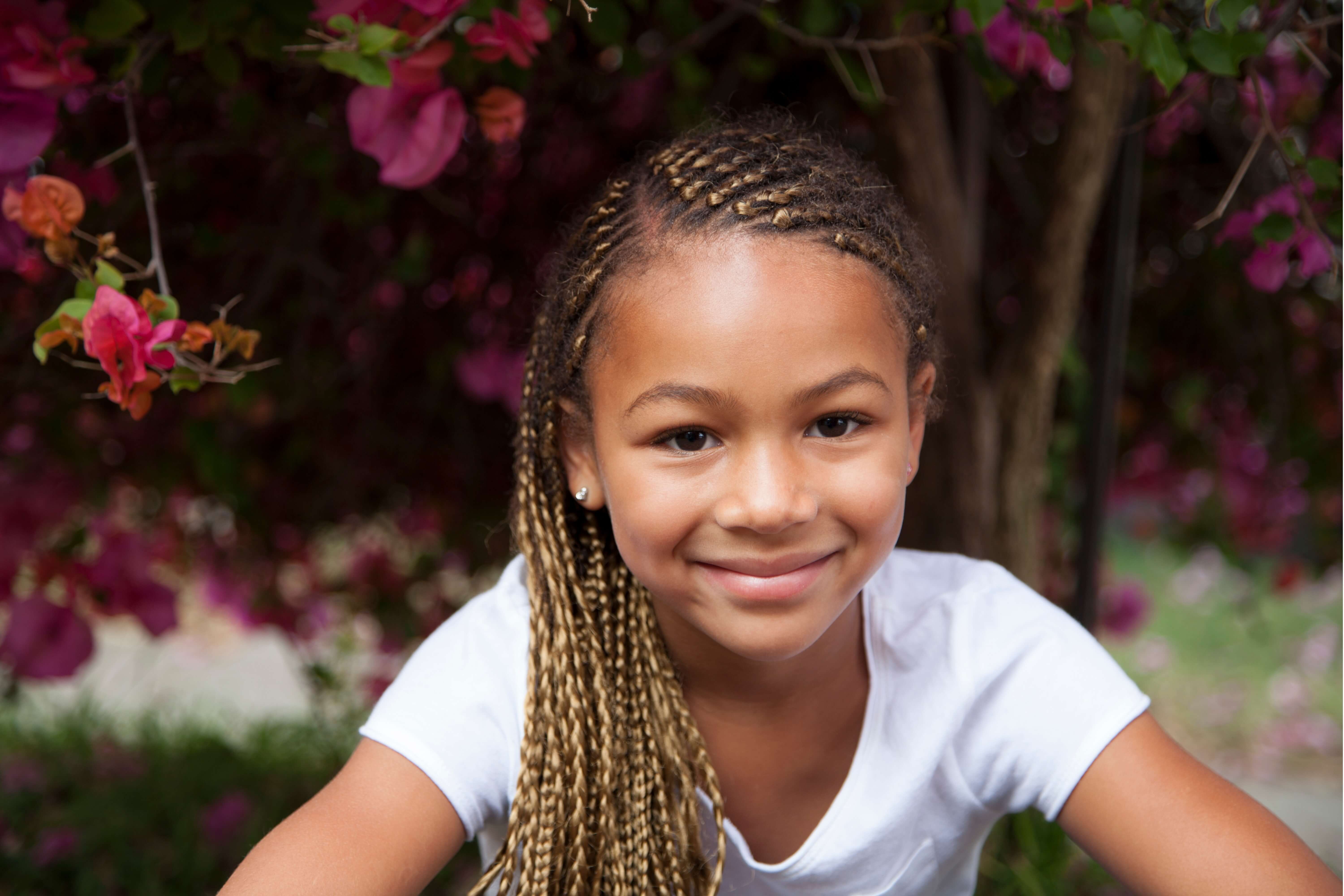 Smiling young girl with braided hair wearing a white shirt outdoors with pink flowers in the background.