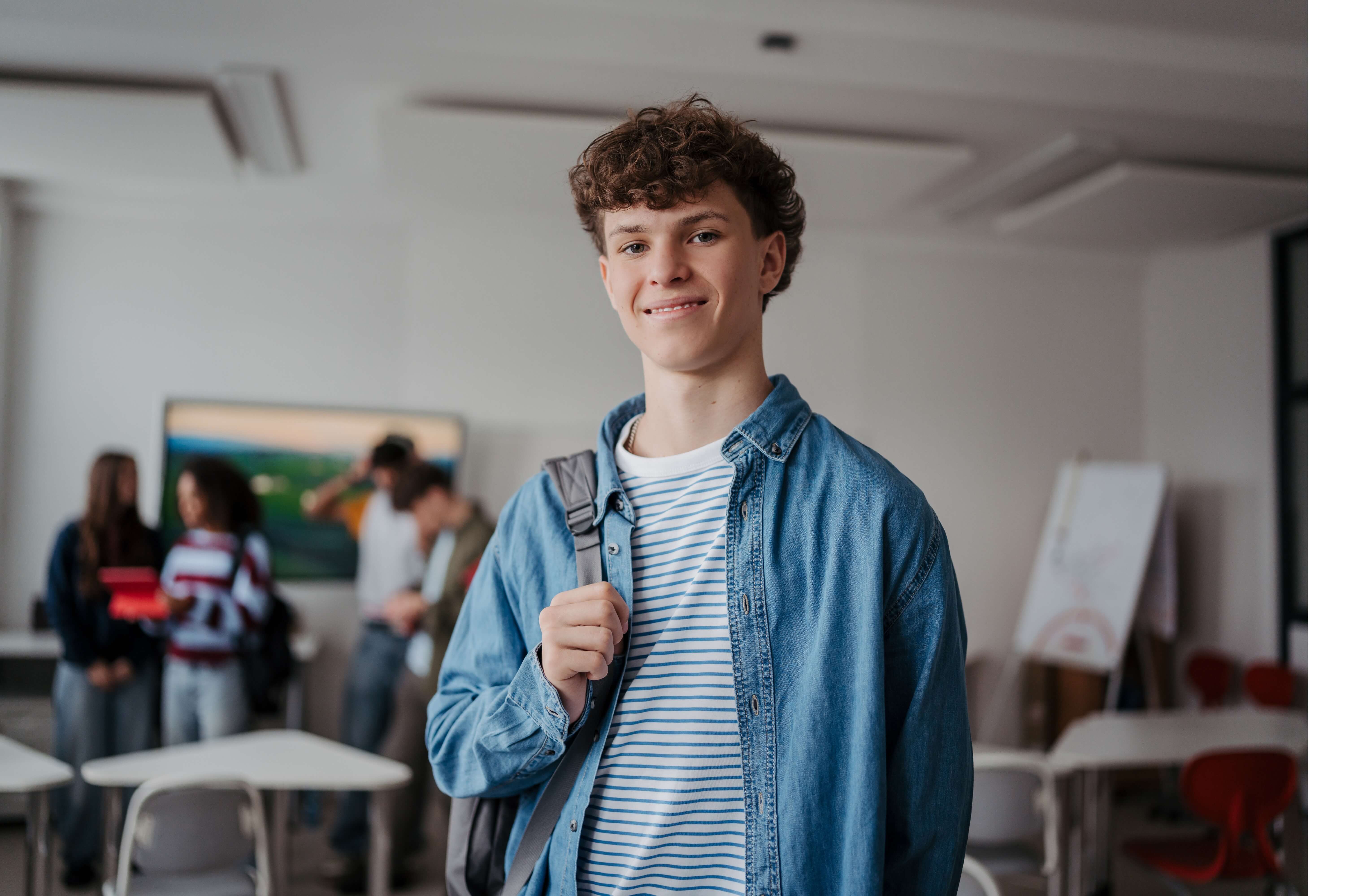 Teenage boy in denim jacket with backpack smiling in classroom with students in background.