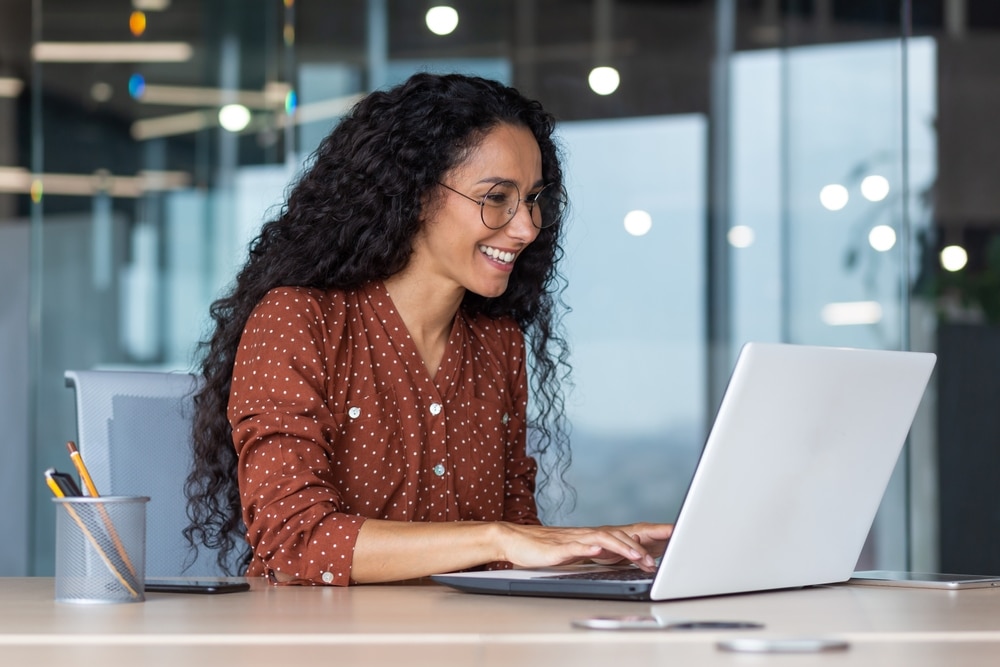 Smiling woman with curly hair and glasses typing on a laptop at a desk in an office.