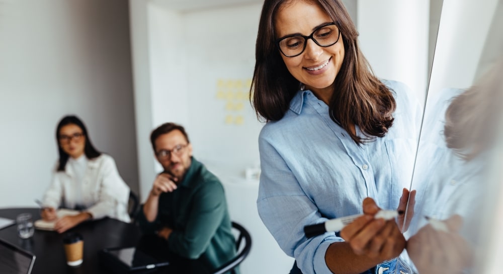 Woman writing on a whiteboard while two colleagues watch attentively in a modern office.