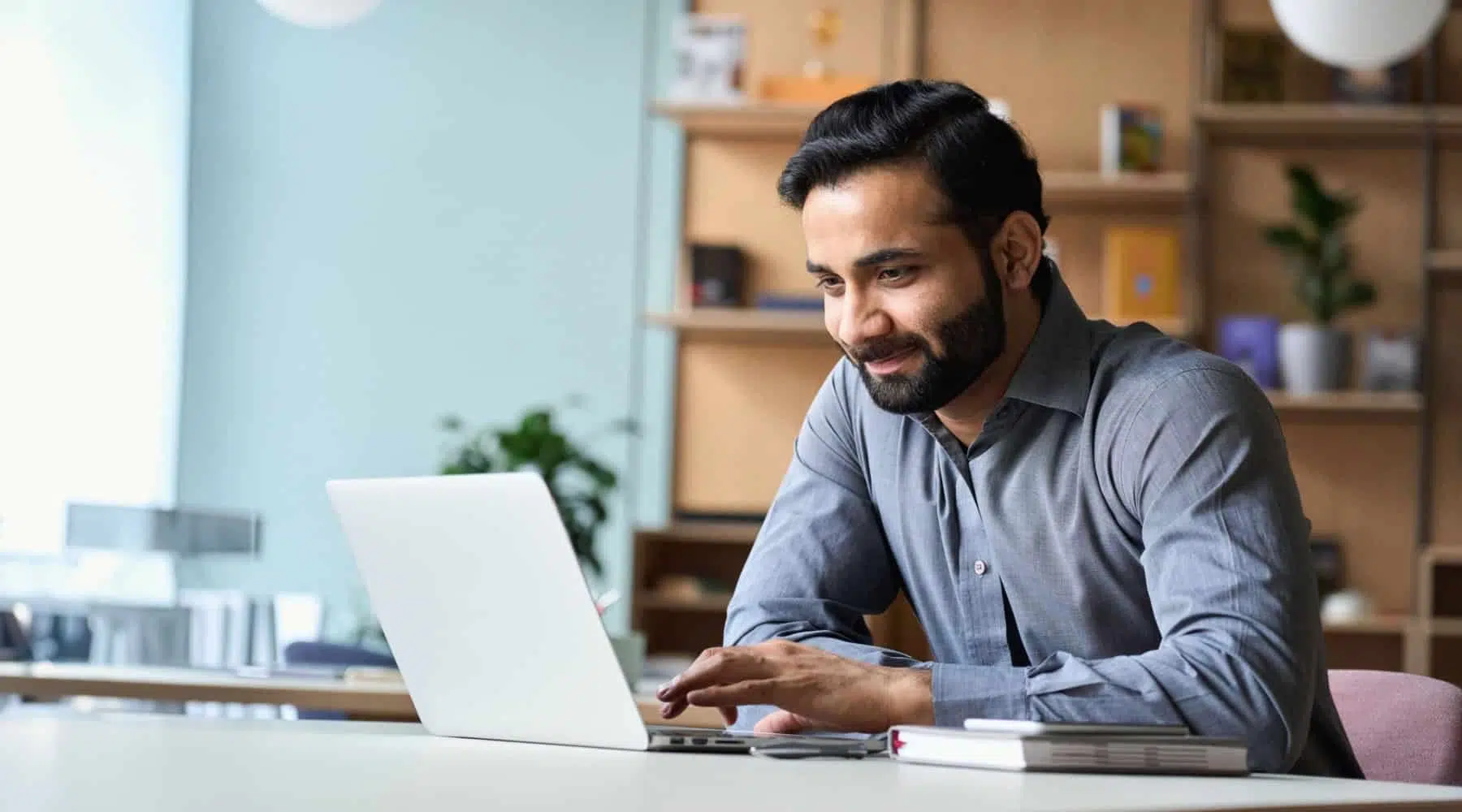 Man smiling while working on a laptop at a desk with a notebook and phone nearby.