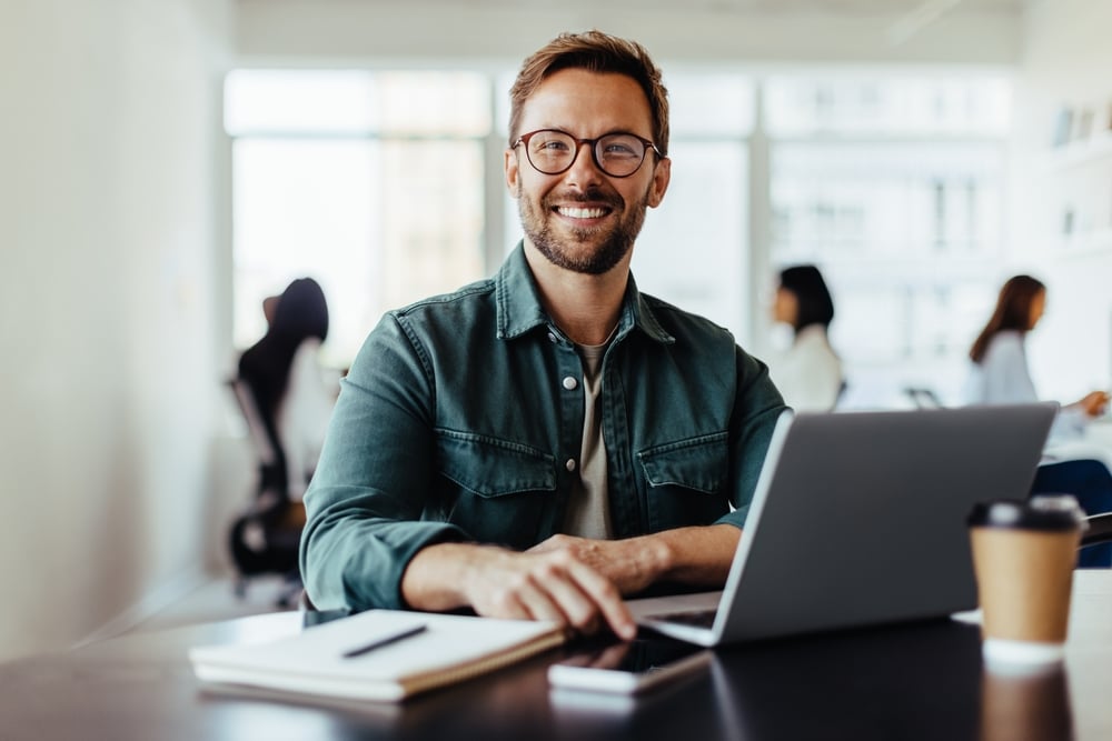 Smiling man with glasses sitting at a desk with laptop, notebook, and coffee in a bright office.