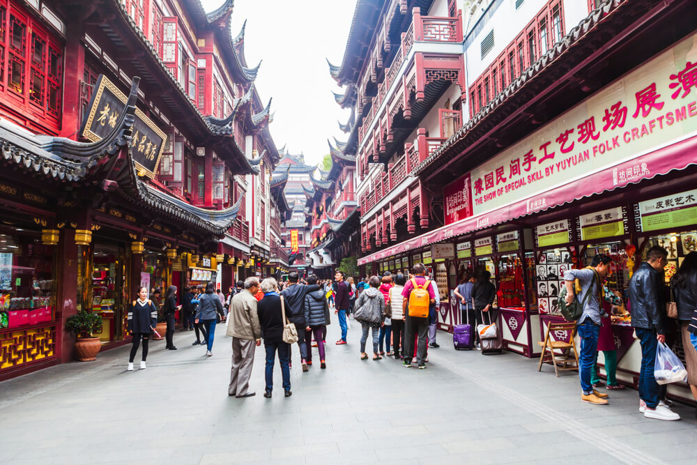 People walk and shop along a busy street lined with traditional Chinese architecture and market stalls.