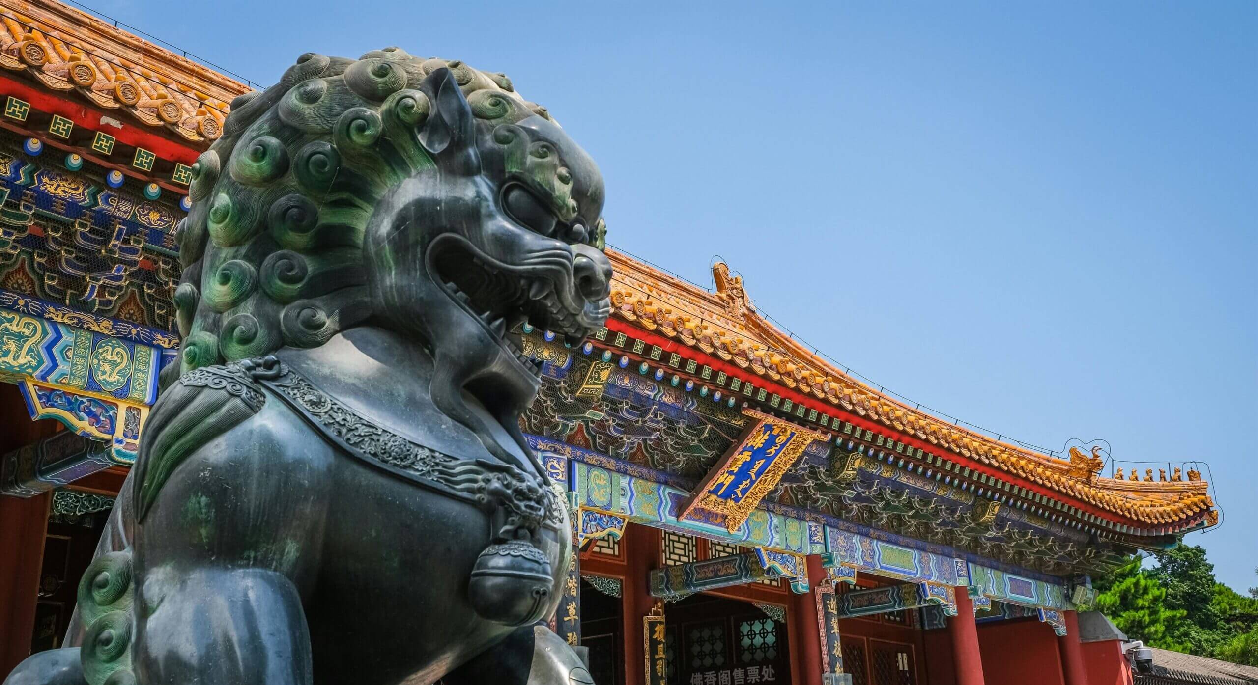 Large stone Chinese guardian lion statue in front of a colorful traditional temple under blue sky.