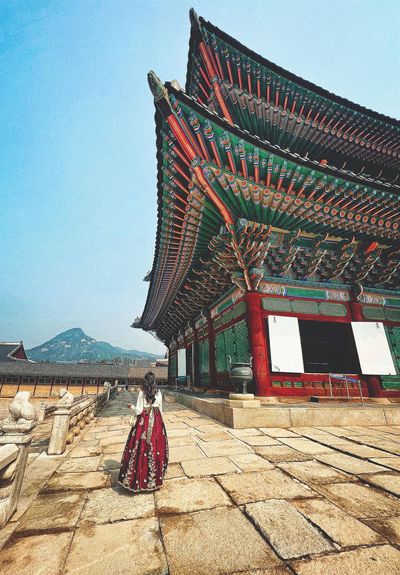 A woman in a traditional Korean hanbok walking near a colorful palace building with a mountain in the background.