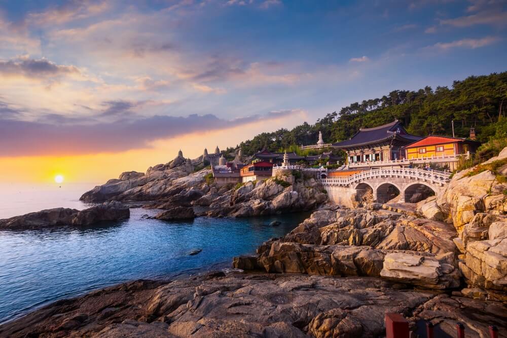 Sunset over rocky coastline with traditional temple buildings and a white arched bridge.