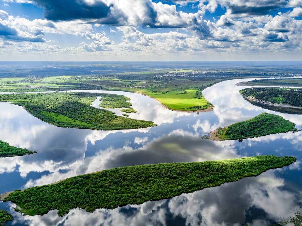 Aerial view of a winding river with green islands and clouds reflected on the water surface.