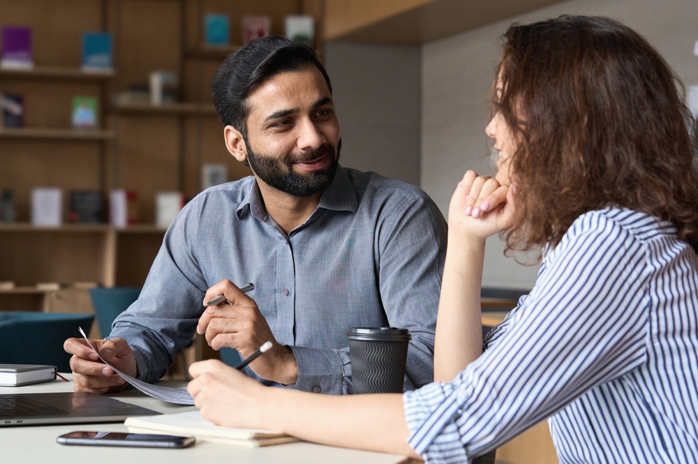 Two people engaged in conversation at a table with a laptop, coffee cup, and notebook.