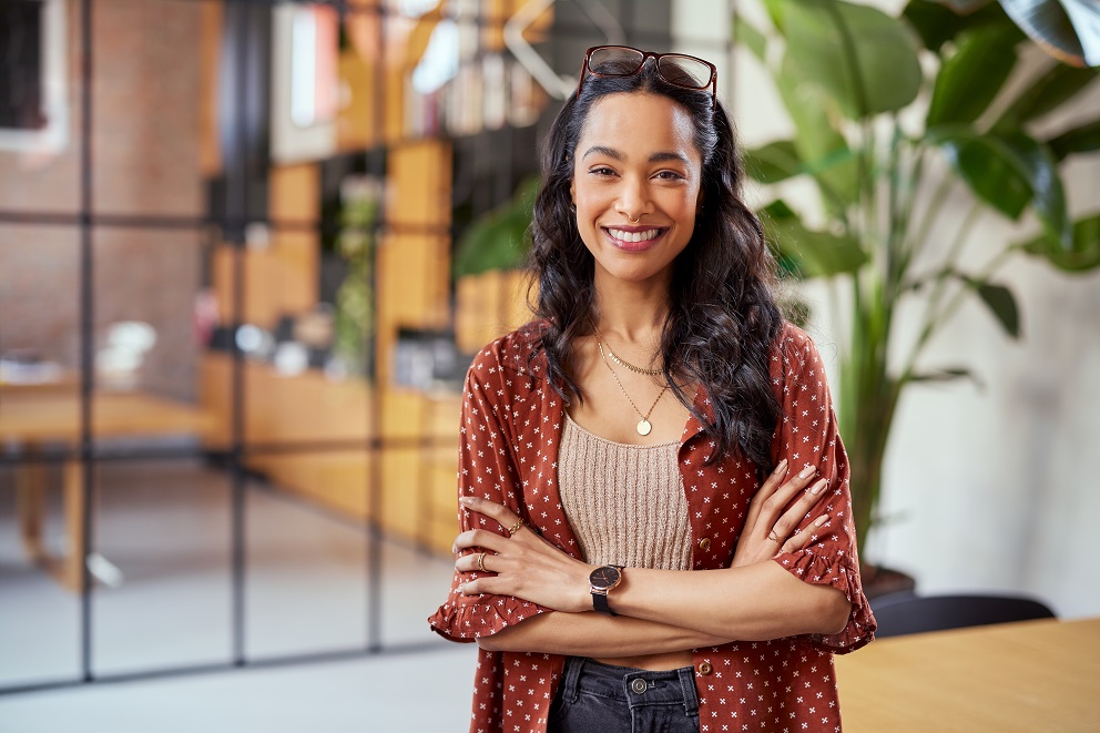 Smiling woman with glasses on her head and arms crossed standing in a modern office space with plants.