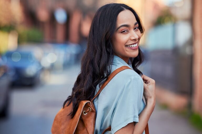 Smiling woman with long black hair wearing a light blue shirt and brown backpack outdoors.