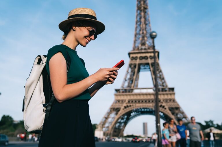Woman with hat and backpack smiling at phone near the Eiffel Tower in Paris on a sunny day.
