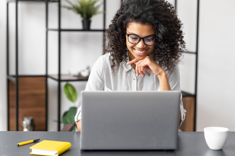 Smiling woman with curly hair and glasses working on a laptop at a desk with a yellow notebook and coffee cup.