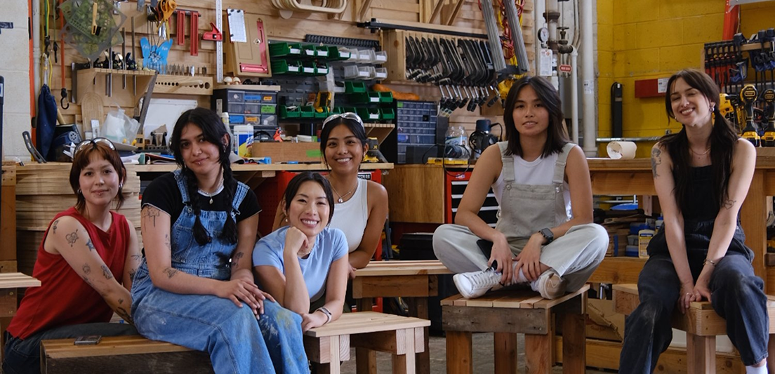 Six women sitting and smiling in a workshop filled with tools and wooden furniture.