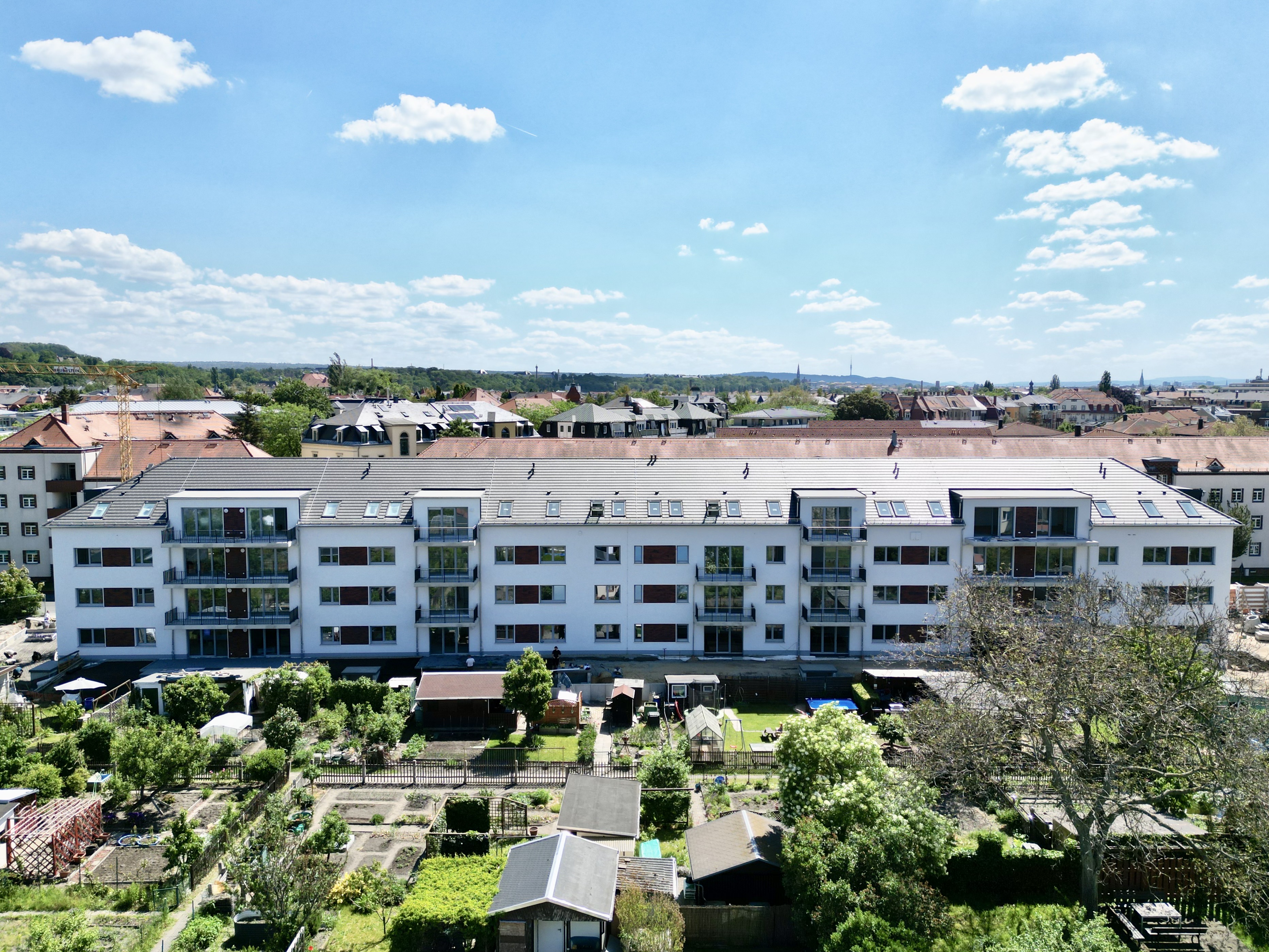 Modern white apartment building with balconies, surrounded by green gardens and small sheds under a partly cloudy blue sky.