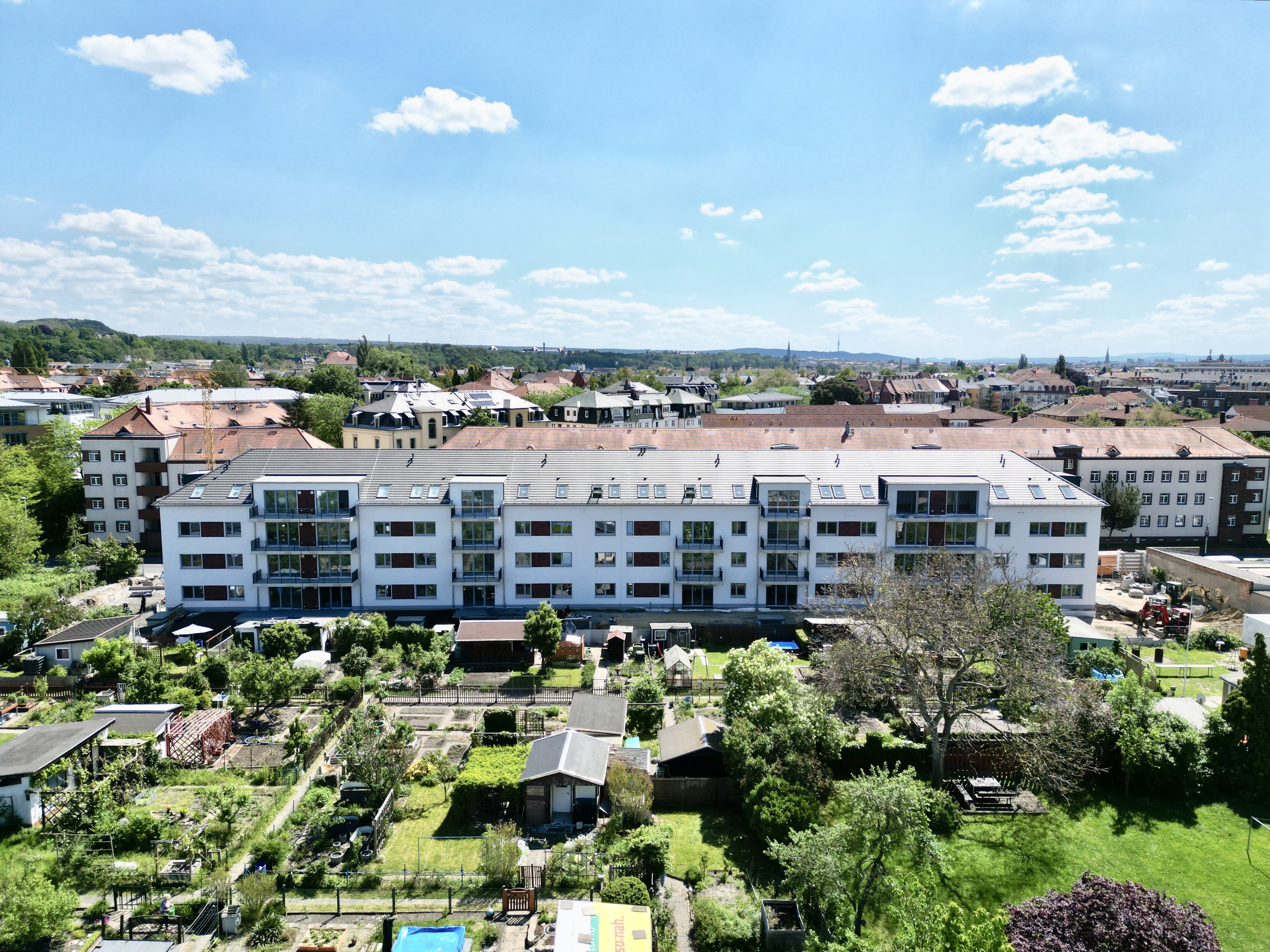 Aerial view of a white residential building with balconies, surrounded by green community gardens and trees under a blue sky with scattered clouds.