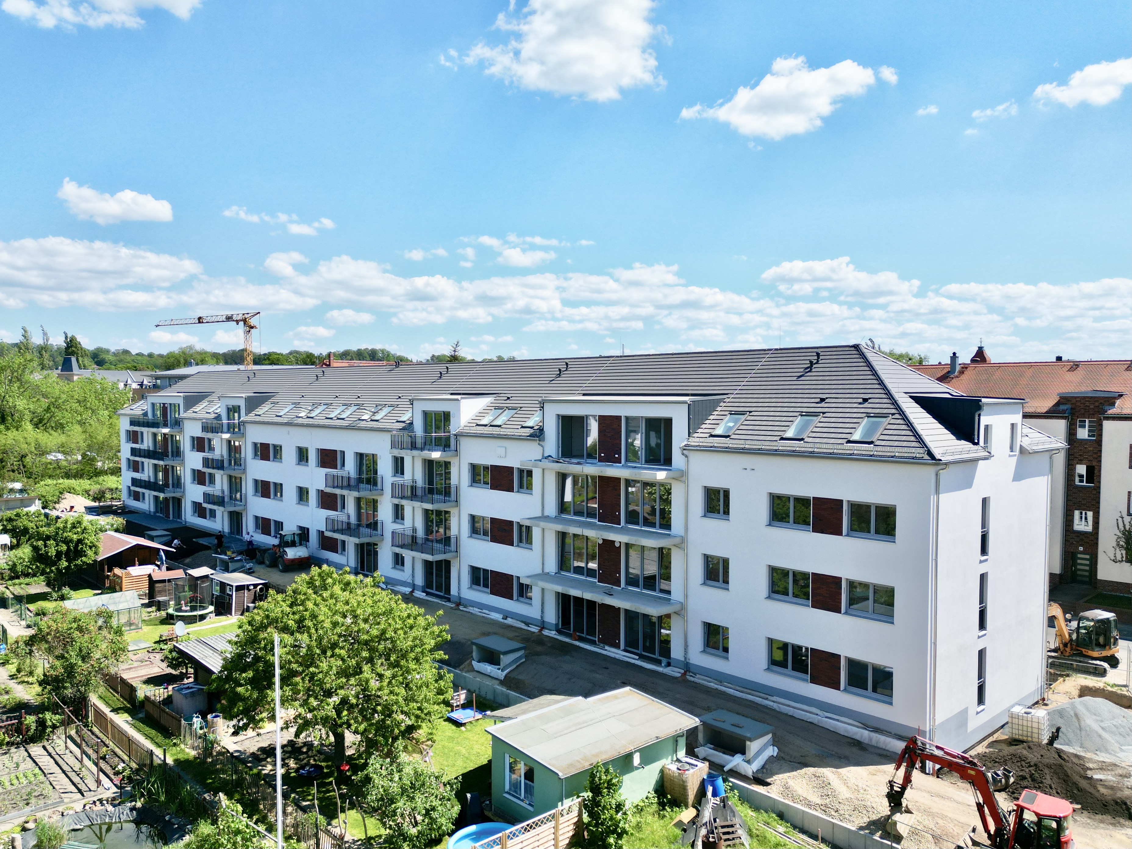 Modern white apartment building with multiple balconies under a blue sky, surrounded by greenery and construction equipment.