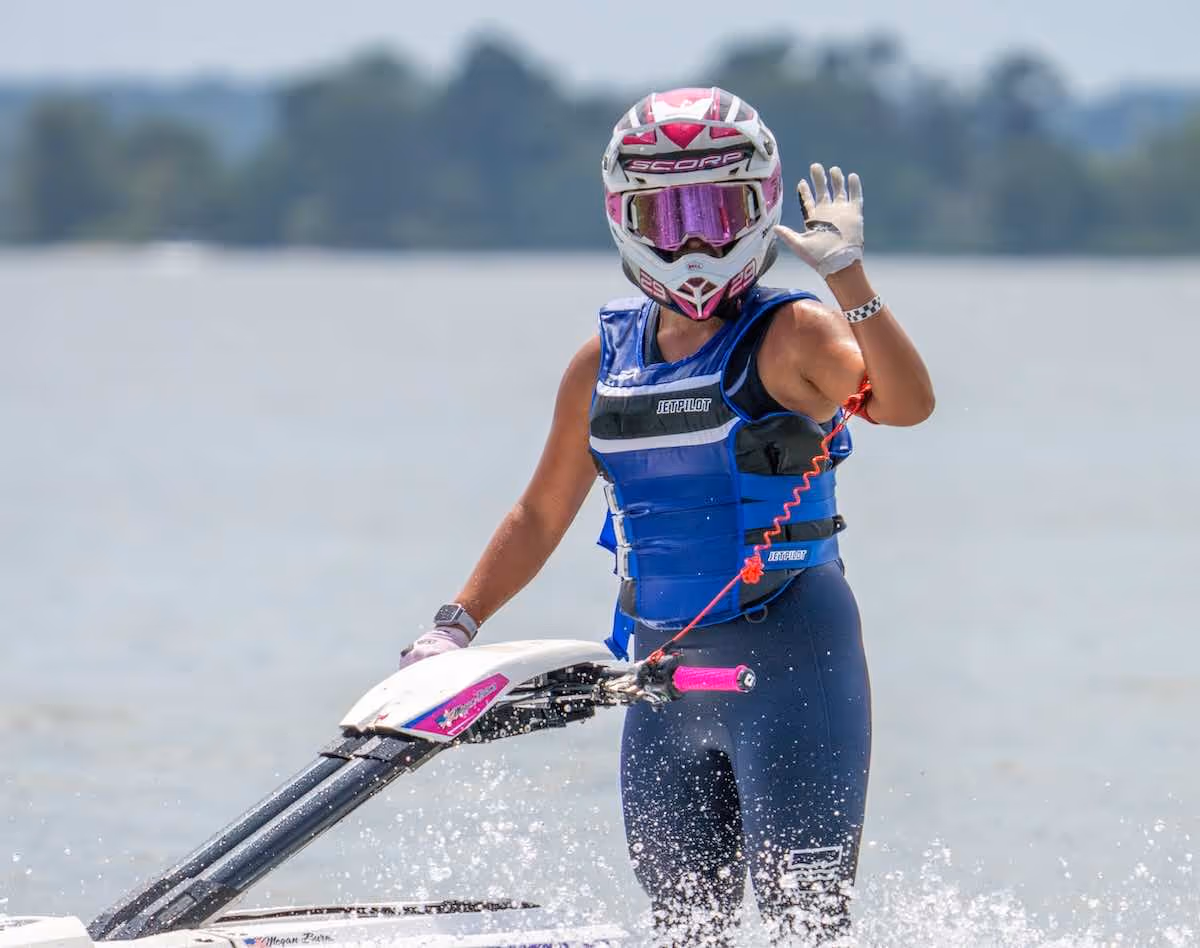 Person wearing a helmet and life vest, waving while standing on a watercraft on a lake.