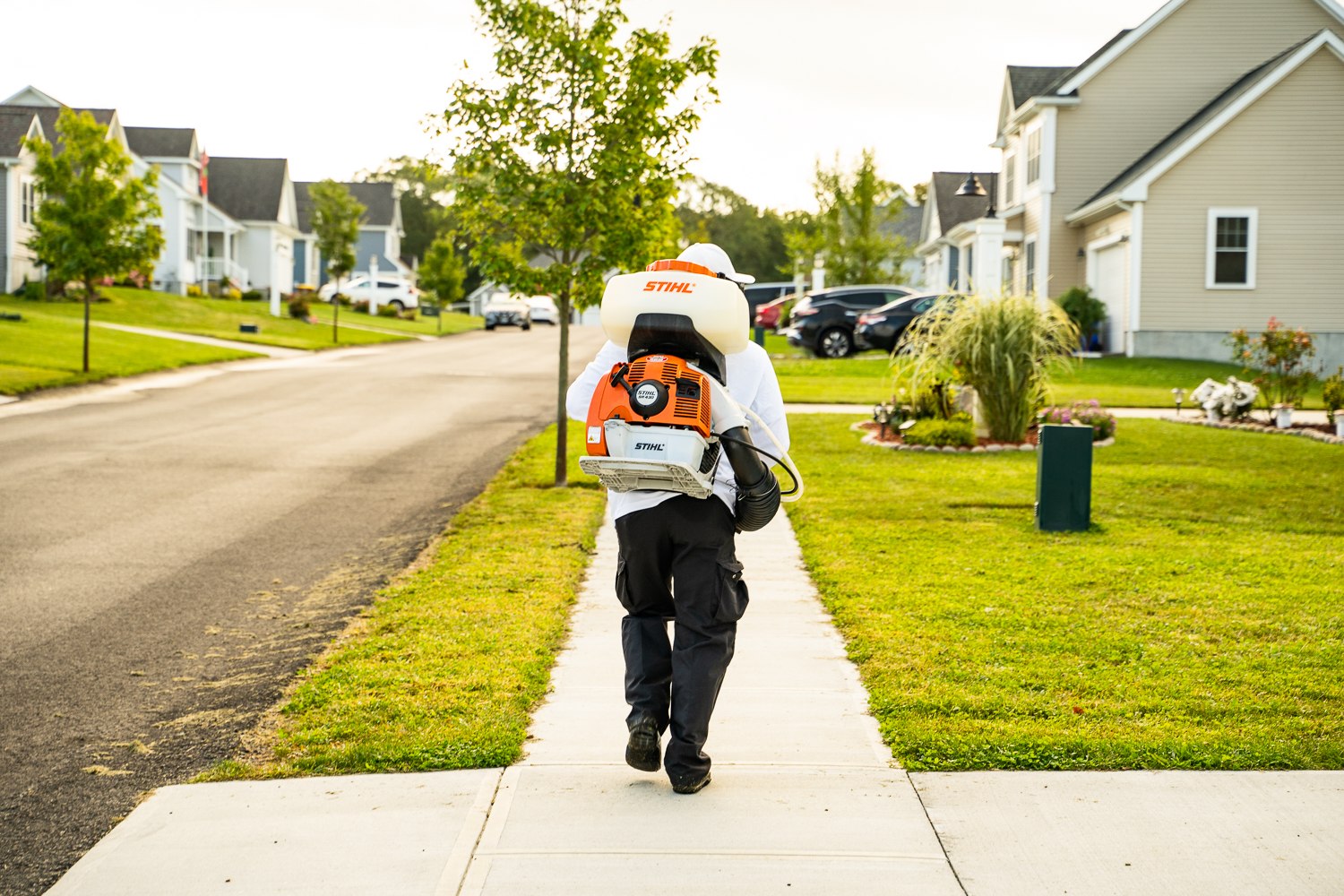 Mosquito Mike technician with a backpack mister walking a neighborhood sidewalk between front yards
