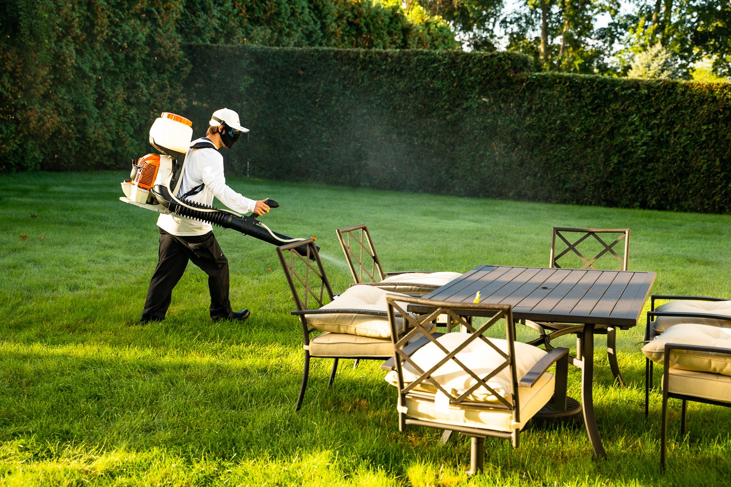 Mosquito Mike technician with a backpack mist blower treating a backyard beside a patio dining set