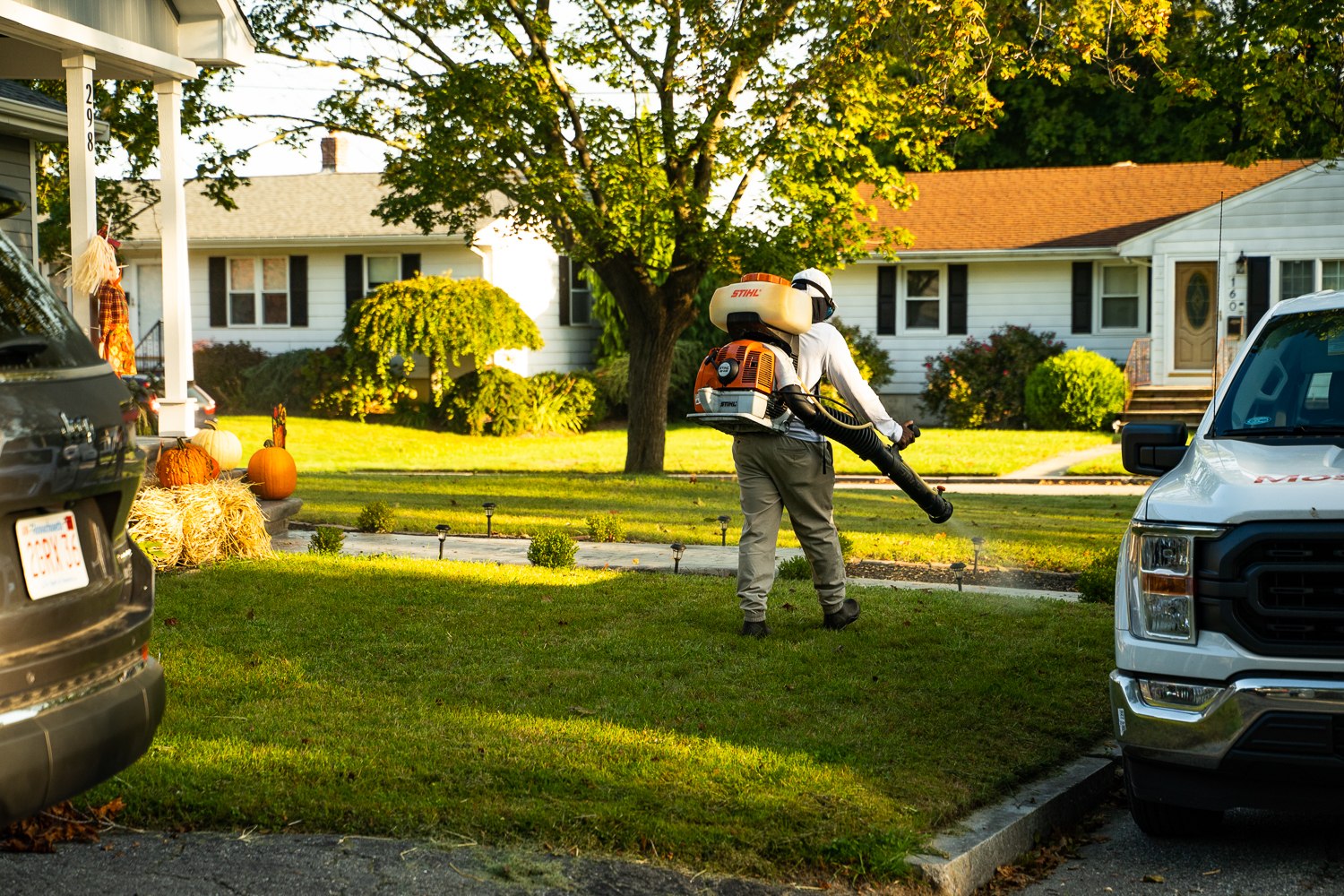Mosquito Mike technician with a backpack sprayer crossing a front lawn on a fall afternoon, with pumpkins and hay bales on the porch steps