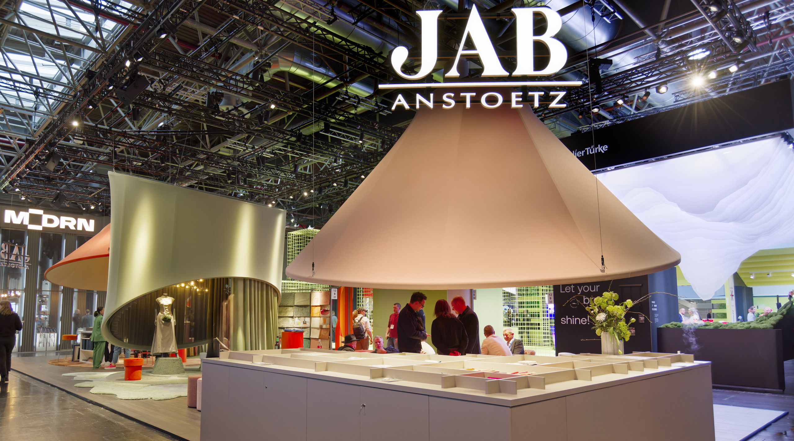 Messestand von JAB Anstoetz mit großen, konischen Hängelampen und Personen, die sich unterhalten.