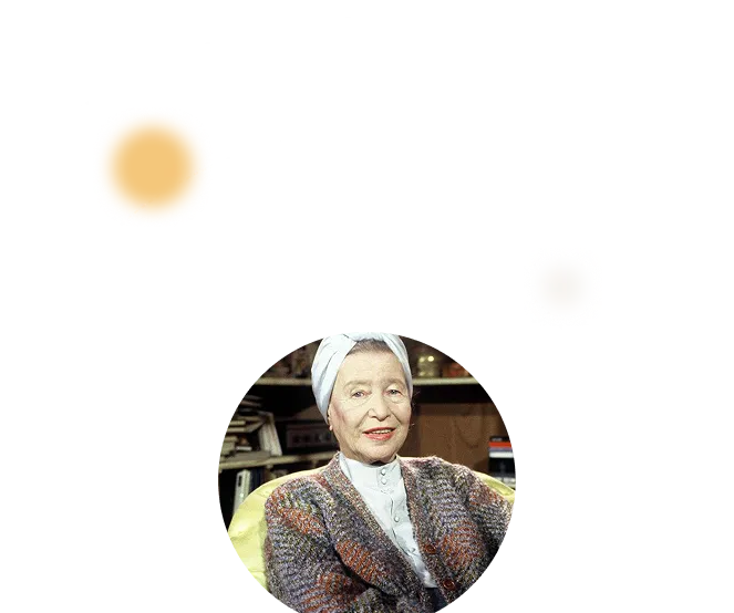 Portrait of an elderly woman wearing a white headscarf and a colorful knitted cardigan, sitting indoors with books and shelves in the background.