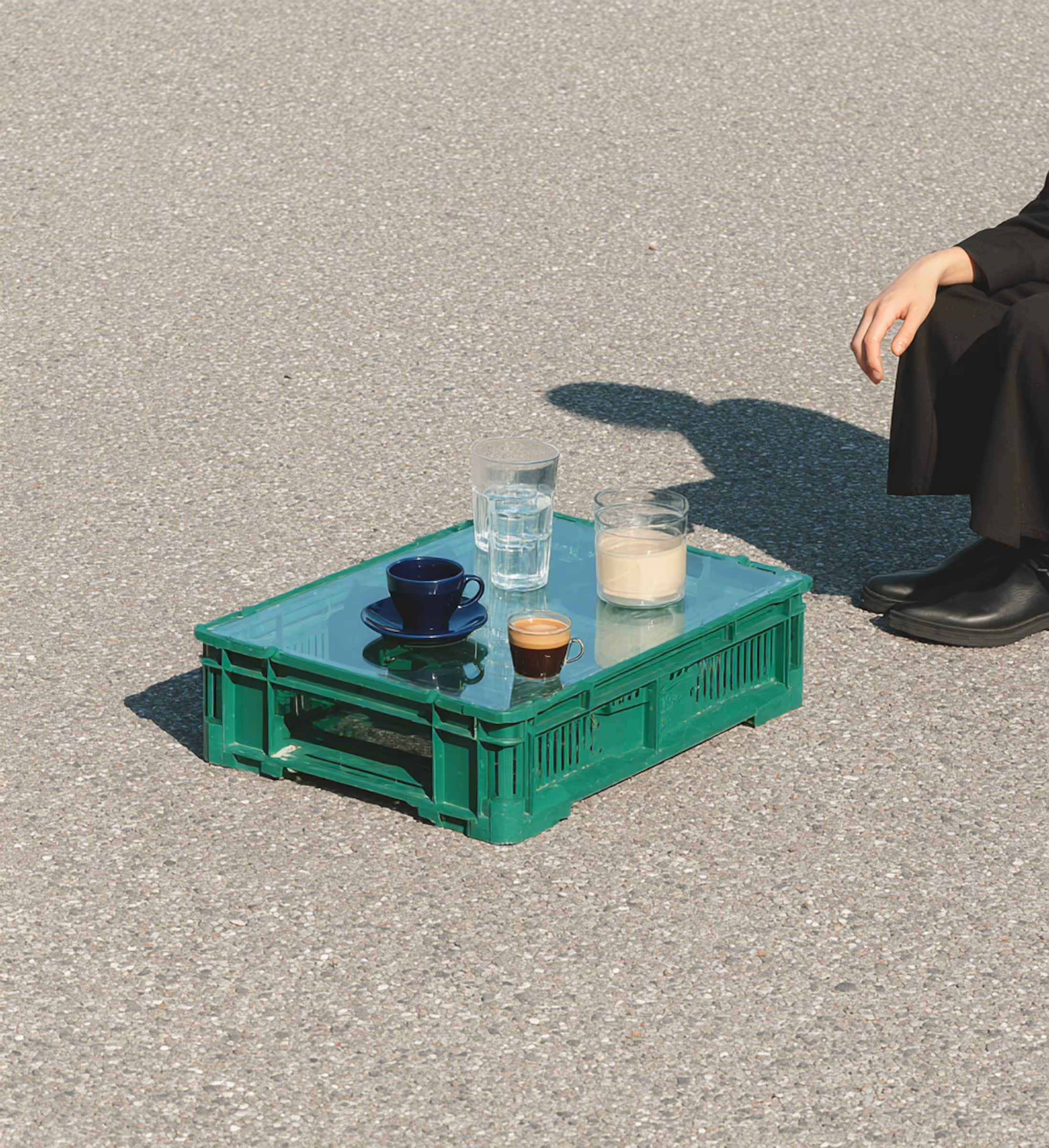 Green plastic crate used as a low table on asphalt with a coffee cup, espresso, glass of water, and a glass candle, next to a person dressed in black.