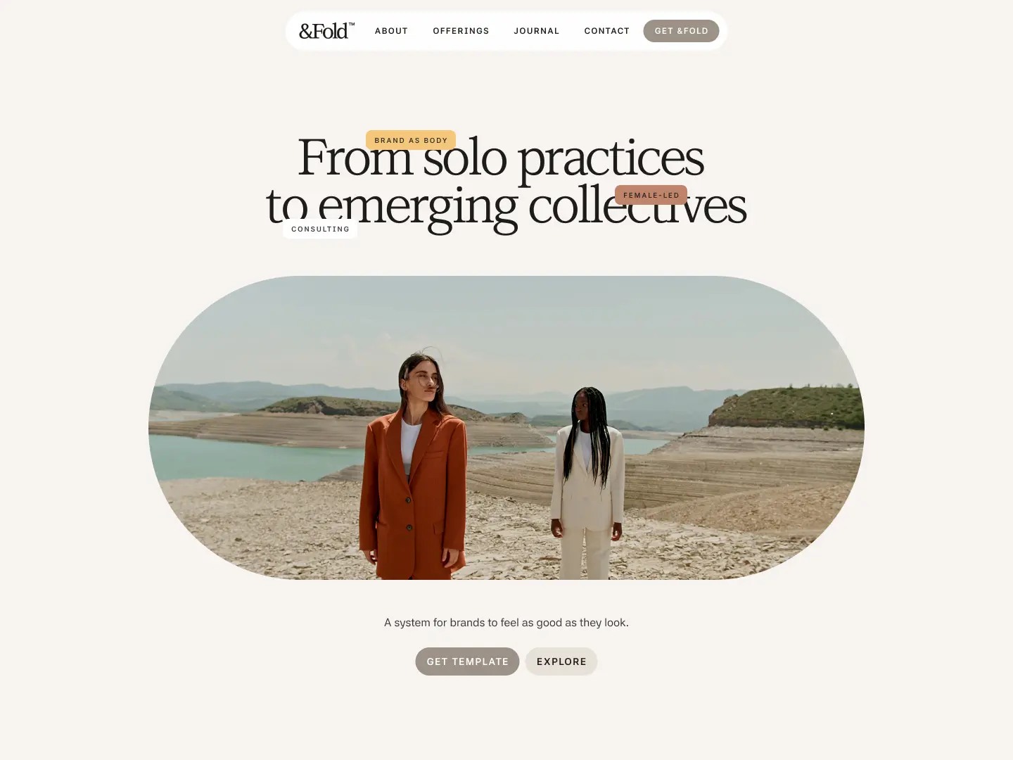 Two women standing outdoors on rocky terrain with water and hills in the background, wearing formal suits in brown and white.