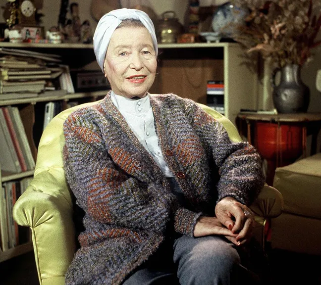 Elderly woman wearing a white headscarf and a multicolored knitted cardigan, sitting in a yellow armchair in a cozy indoor setting with books and decorative items in the background.