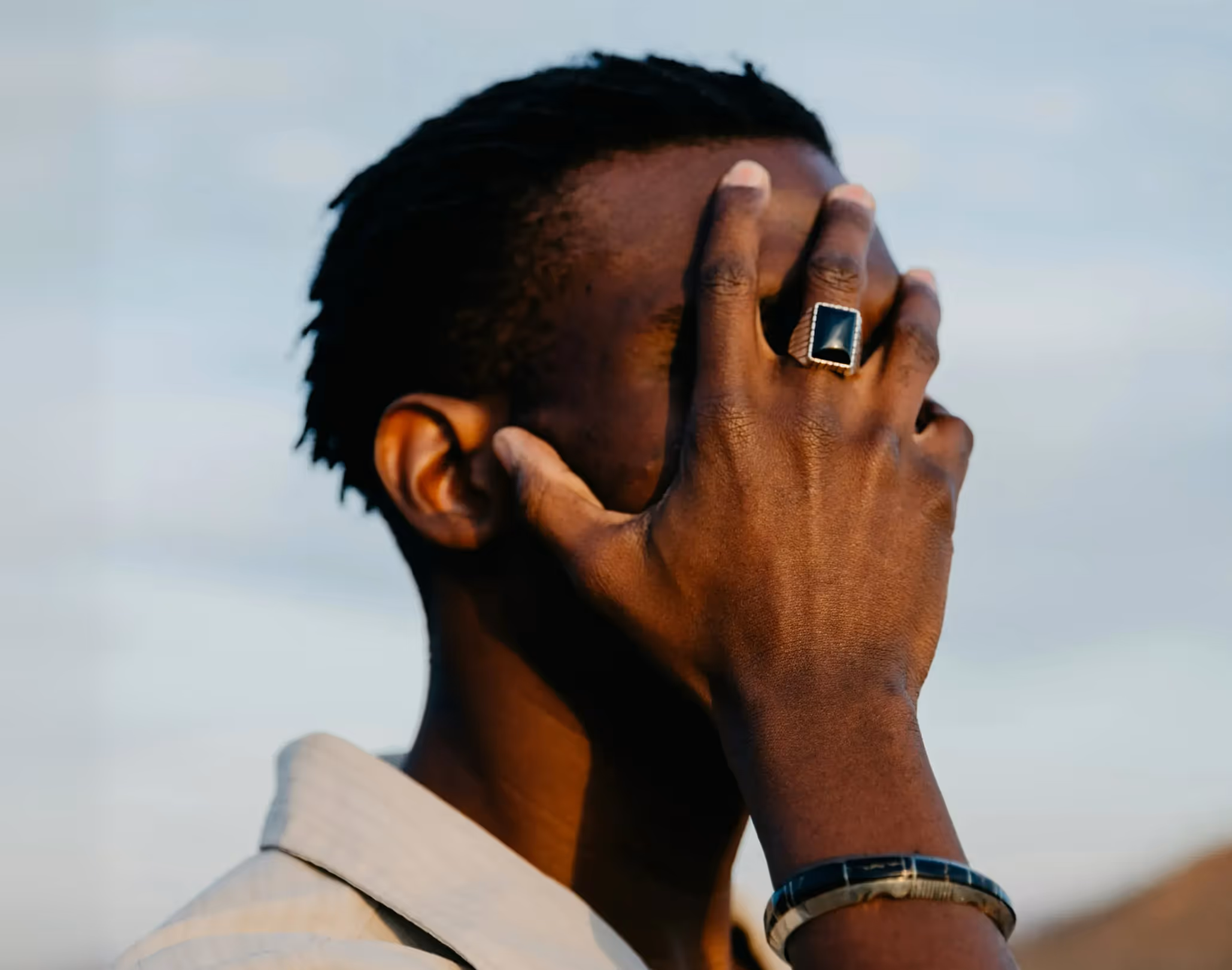 Man with dark skin covering his face with his hand, wearing a large square black ring and a wristwatch.