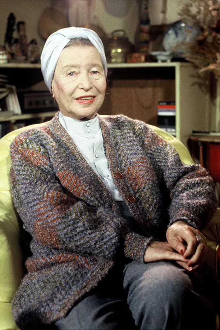 Elderly woman wearing a patterned cardigan and white headscarf sitting on a yellow armchair, smiling.