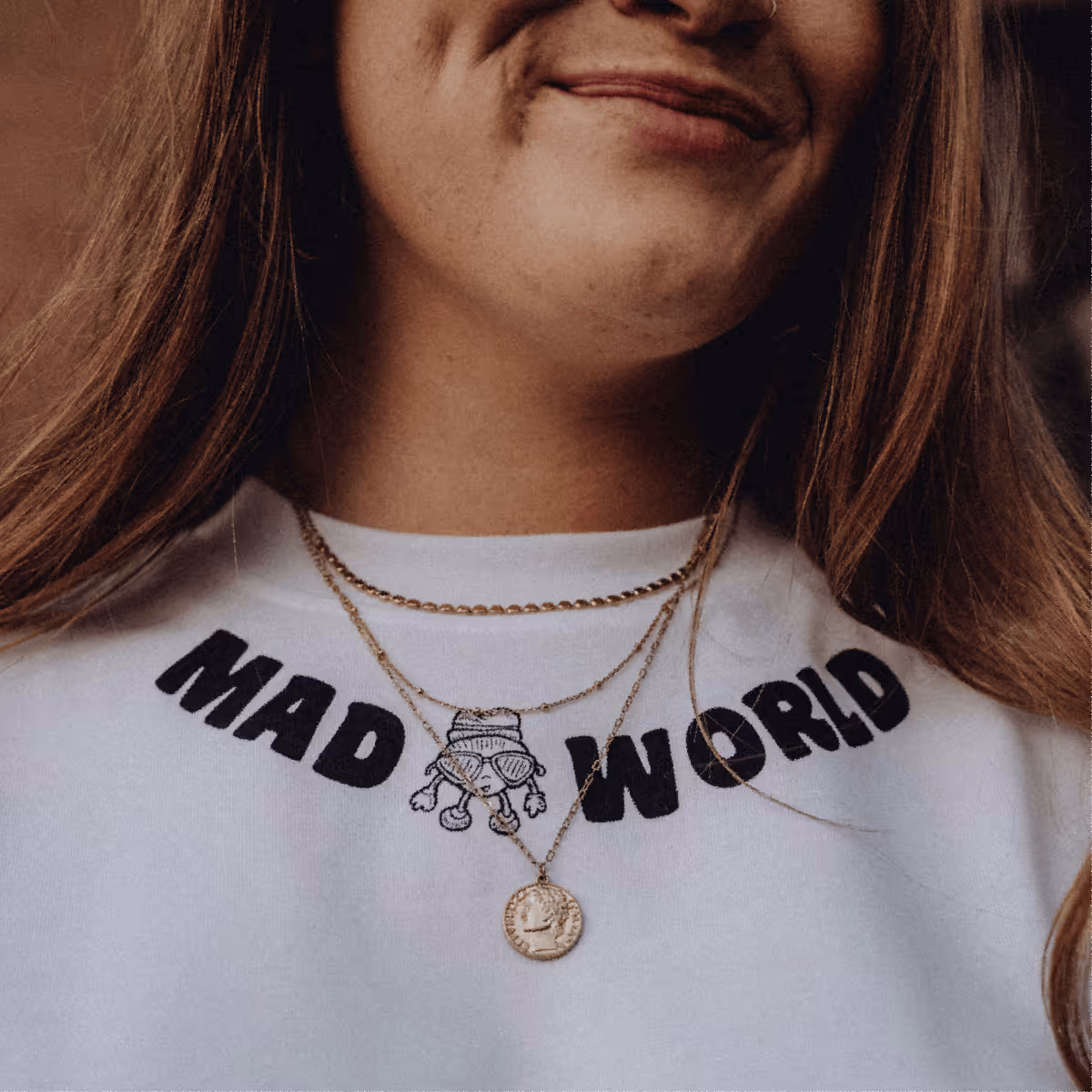 Close-up of a person wearing a white shirt with black text 'MAD WORLD' and a small cartoon character between the words, along with layered gold necklaces.