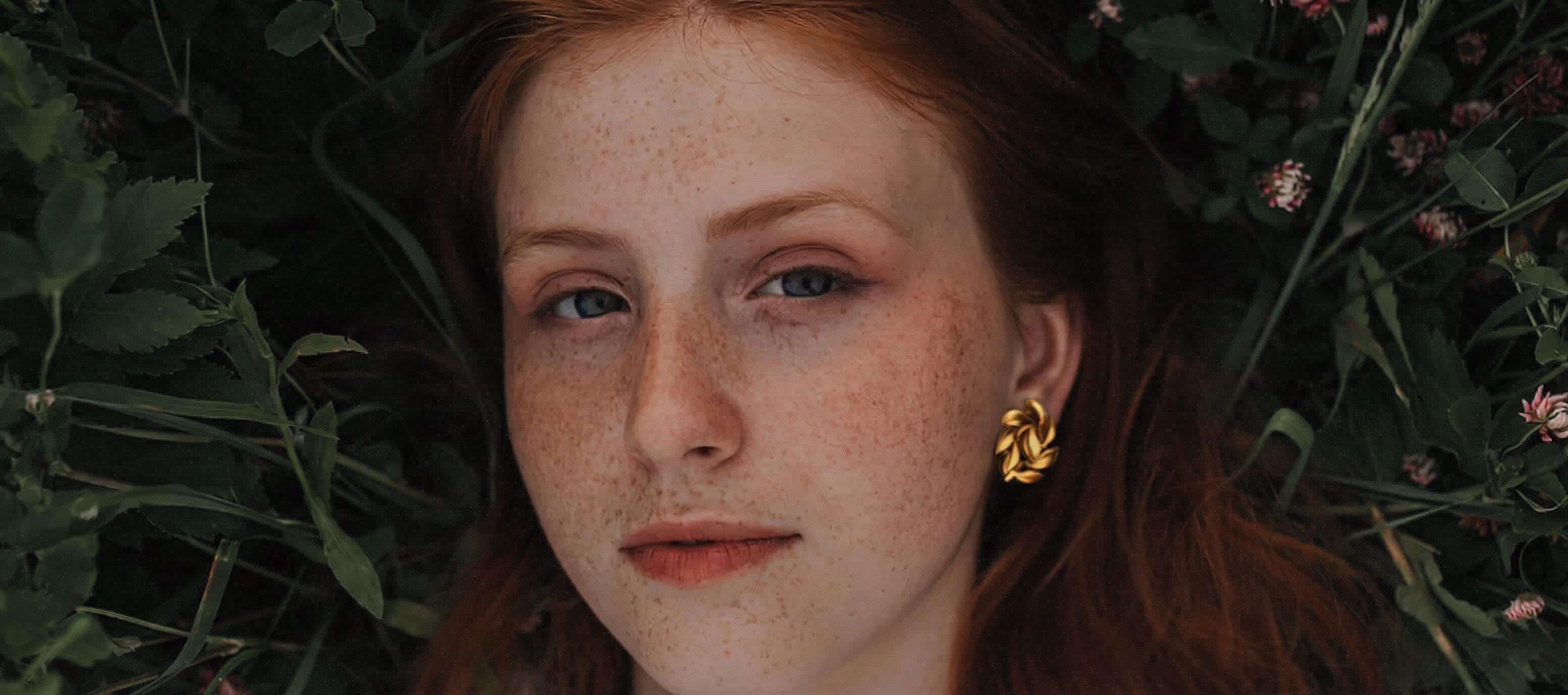 Close-up of a young woman with red hair and green eyes lying on green foliage with small pink flowers.
