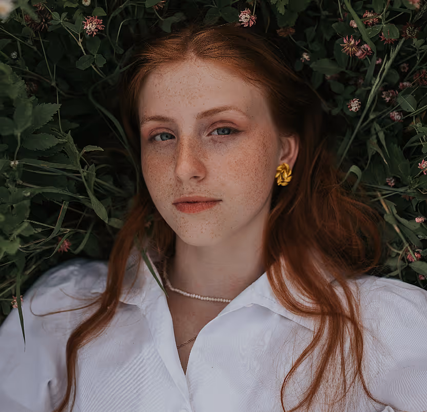 Red-haired woman with freckles lying on grass wearing a white blouse, pearl necklace, and gold earrings.