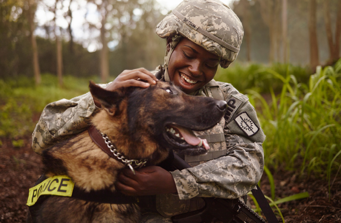 Smiling female soldier in camouflage uniform petting a police dog in a forested area.