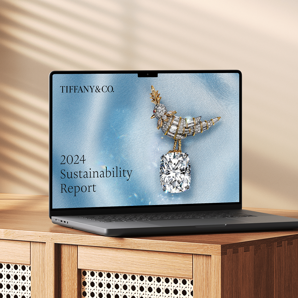 Open laptop on wooden cabinet screen showing Tiffany & Co. 2024 Sustainability Report with a diamond bird brooch on blue background.