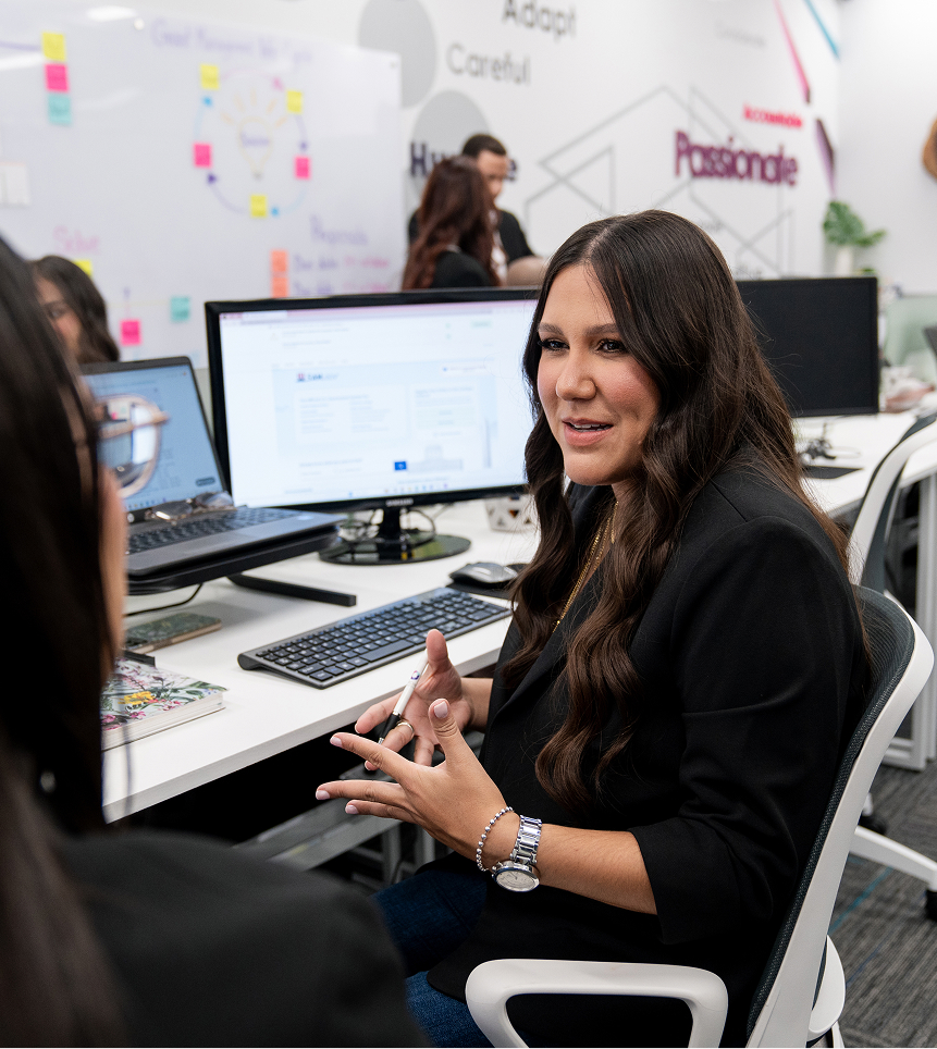 Woman with long dark hair wearing a black blazer talking and gesturing in a modern office with multiple computer workstations.