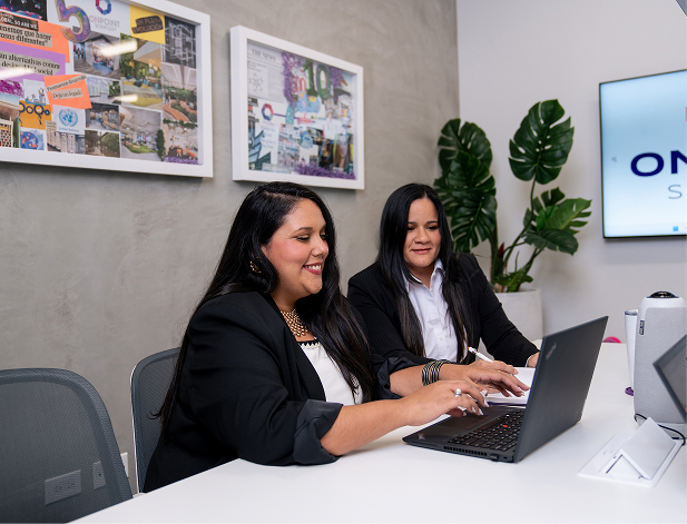 Two women in business attire working together on a laptop in a modern office setting.