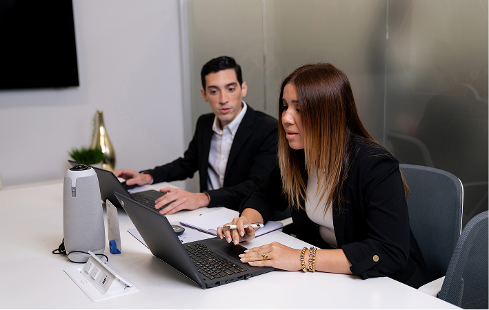Woman in a purple blazer writing on paper at a white desk in a modern office with computer monitors, a printer, and a colorful wall.