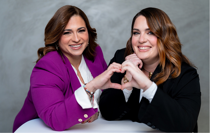Woman in a purple blazer writing in a notebook at a desk with computer monitors and office supplies.
