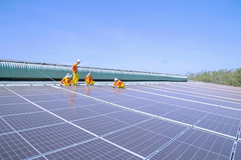 Four workers in orange and yellow uniforms installing solar panels on a large rooftop under a clear blue sky.