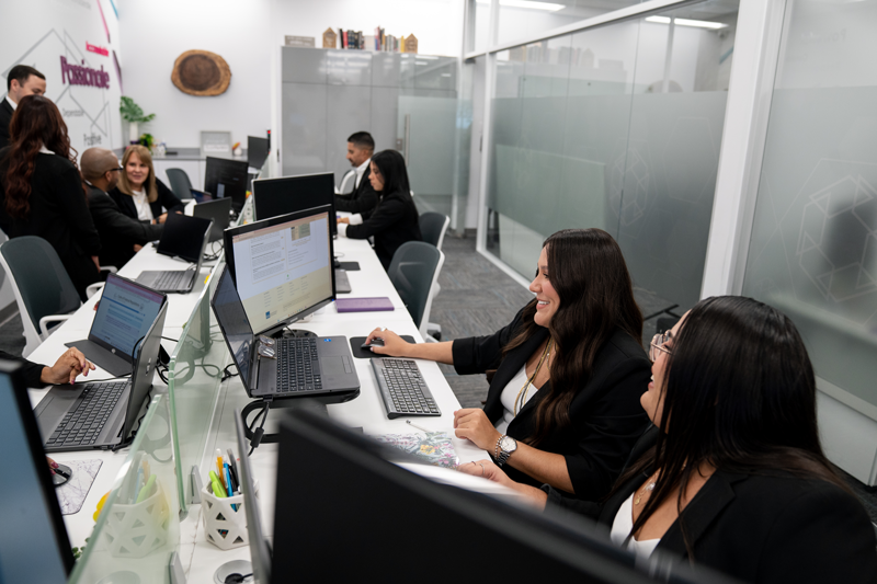 Office scene with multiple people working at desks with laptops and desktop computers, some engaged in conversation.