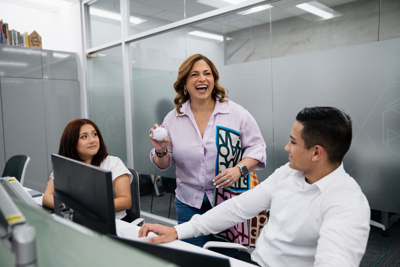 Smiling woman holding a stress ball interacting with two coworkers seated at computers in an office.