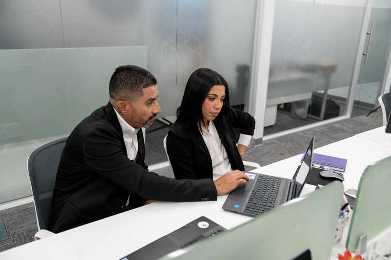 Two business professionals in black suits working together on a laptop at a modern office desk.