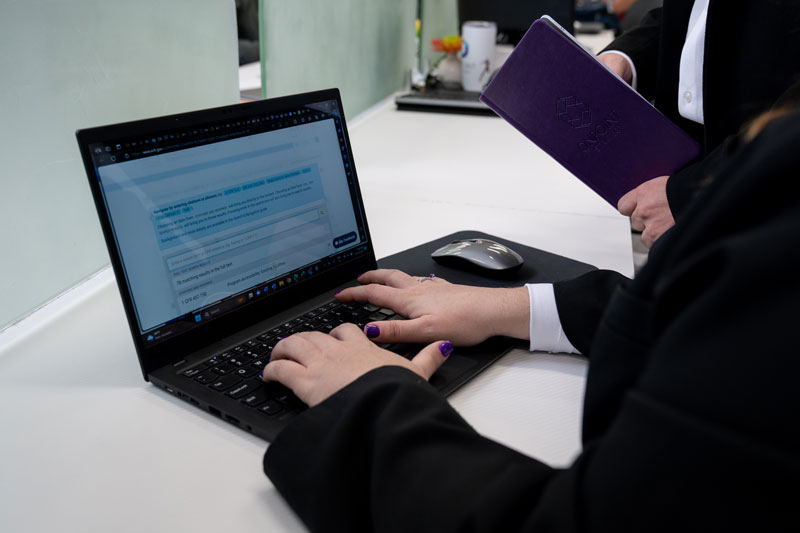 Person with purple-painted nails typing on a laptop keyboard with a wireless mouse nearby and another person holding a purple portfolio standing beside them.