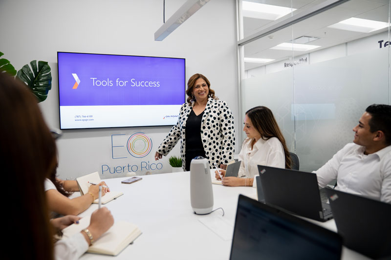 A woman in a white and black polka dot blazer leading a business meeting with three attendees seated around a table with laptops and notebooks, with a screen displaying 'Tools for Success' in the background.